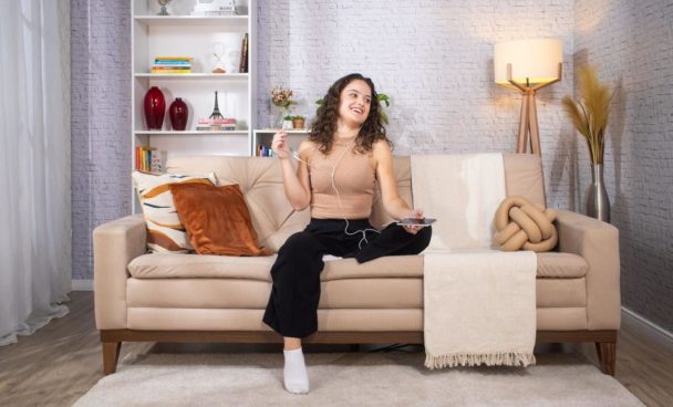 : uma mulher branca de cabelo castanho veste roupas confortáveis nas cores bege e preto, assim como uma meia. Ela está sentada em um sofá bege, ambientado em uma sala de estar. Em cima do sofá estão almofadas em diversas texturas e tonalidades e, ao fundo, estantes com objetos decorativos e luminárias decoram o ambiente.