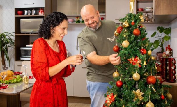 Casal de adultos, decorando juntos uma árvore de Natal, sorrindo. Ao fundo, uma cozinha Madesa e uma mesa de jantar, ambos decorados.