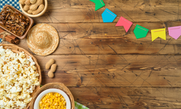 A imagem ilustra o que tem na festa junina em cima de uma mesa de madeira com bandeirinhas coloridas.
