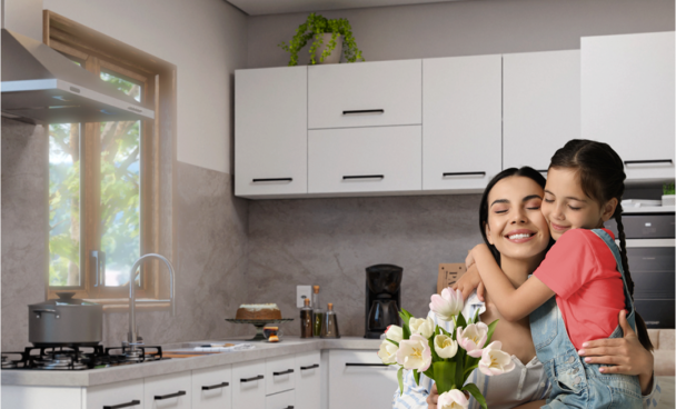 Texto alternativo: mãe sorridente segurando um buquê de tulipas brancas em uma cozinha moderna. Uma menina está abraçando a mãe, sorrindo. A cozinha tem armários brancos e uma janela que deixa entrar luz natural. Um cartão decorado está sobre a mesa.