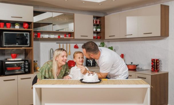 Família feliz, com a mãe, pai e filho sorrindo em frente a um bolo de aniversário branco. Ao fundo, uma cozinha Madesa na cor crema, decorada com utensílios vermelhos.