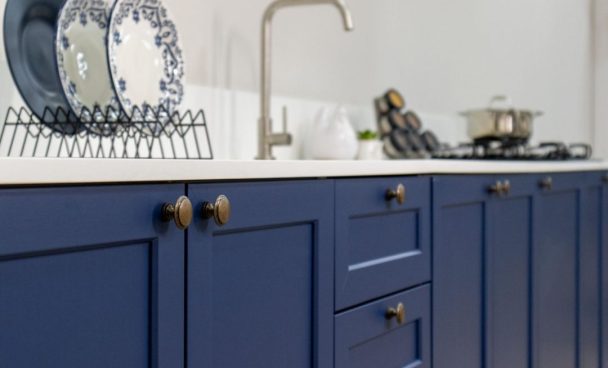 Armário de cozinha azul com portas e gavetas, com detalhes em maçanetas douradas, e pia com torneira ao fundo. Ao lado, utensílios de cozinha, como pratos decorativos brancos com detalhes azuis em um escorredor e uma panela no fogão.