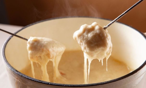 Duas bolas de queijo derretido sendo retiradas de um aparelho de fondue com garfos, com o queijo derretido e borbulhante escorrendo.