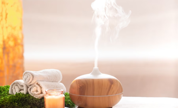 A imagem ilustra o que é aromaterapia através de um difusor de ambientes ligado. Ao lado dele encontra-se uma vela e três toalhas brancas enroladas.