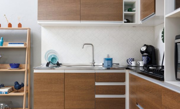 Cozinha Madesa com portas e gavetas em madeira e detalhes em metal está compondo uma cozinha em L, com uma cuba de pia, torneira, e eletrodomésticos sobre sua estrutura.