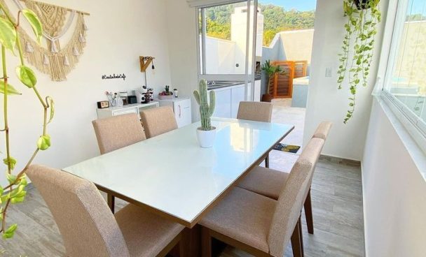 Mesa de jantar branca retangular com seis cadeiras bege, plantas em vasos no centro e ao redor, janelas grandes permitindo entrada de luz natural, e decoração moderna no ambiente.