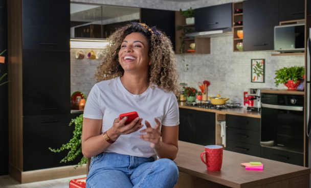 Mulher negra, sorrindo, com o celular em mãos. Ao fundo, está a cozinha Ágata Madesa, na cor preta.