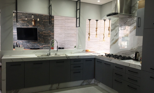 A imagem ilustra uma cozinha americana planejada na cor cinza escuro com uma pedra de mármore branco em cima e detalhes metalizados e pretos, como a pia e a geladeira.