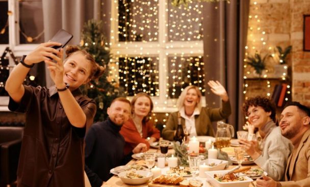 família reunida em mesa de ceia de natal. Menina segura celular e tira selfie com as pessoas sentadas na mesa, que sorriem e olham para a foto.