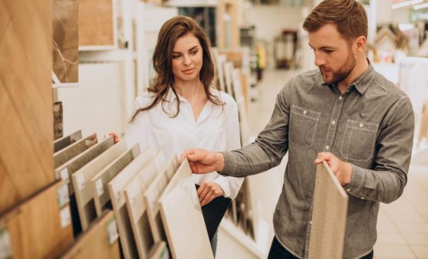 Um homem e uma mulher estão em uma loja de materiais de construção ou decoração, olhando e escolhendo amostras de pisos.