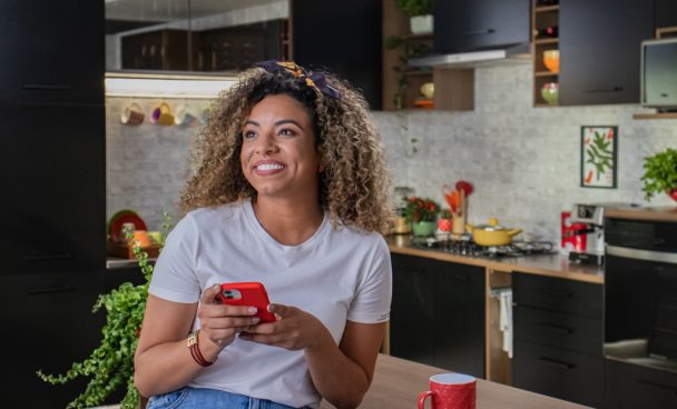 Mulher negra, sorrindo, sentada numa bancada enquanto segura um smartphone.
