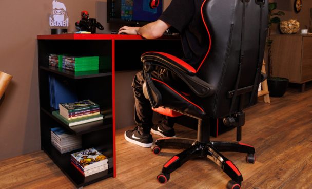 Mesa gamer Madesa, decorada com computador, mouse, livros e itens geek. À frente, cadeira gamer nas cores preto e vermelho.