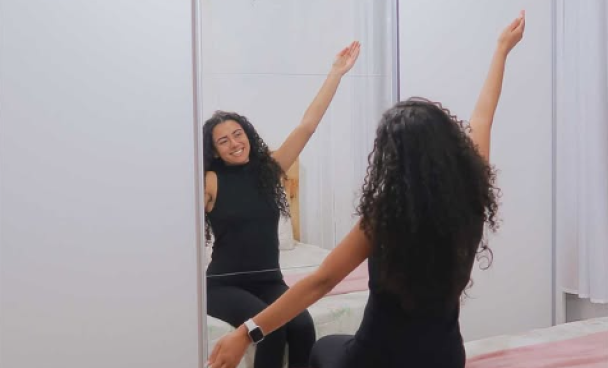 Uma mulher com cabelos cacheados, vestindo roupa preta, sentada na cama de frente para um espelho grande, com os braços levantados e sorrindo.