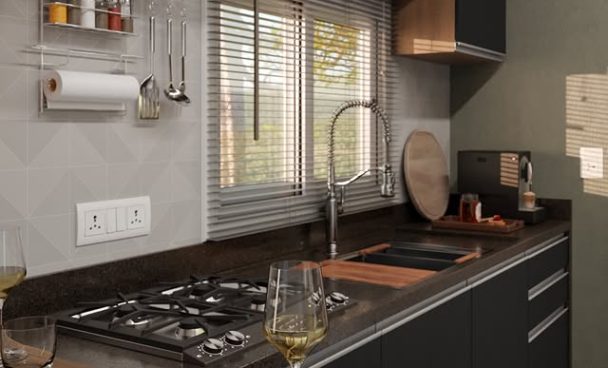 Cozinha com bancadas pretas, janela com persianas, pia com torneira de alta, utensílios pendurados na parede, pratos e copos sobre a mesa de jantar, e uma taça de vinho na frente.