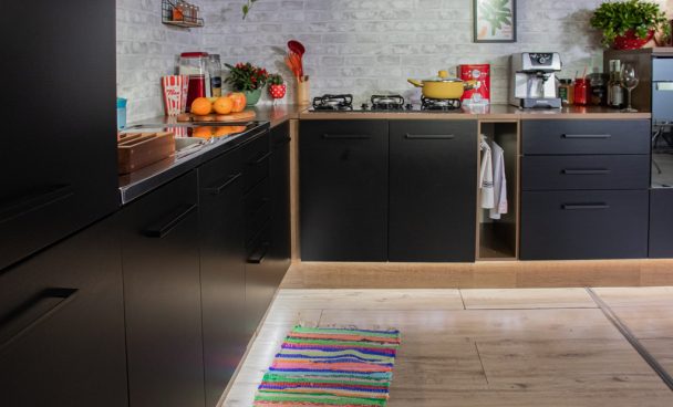 Uma cozinha Madesa Ágata preta está em um ambiente amplo, e acompanha o movimento da parede em L. Há um tapete colorido no chão e, sobre os balcões da cozinha, estão dispostos diversos eletrodomésticos, alimentos e utensílios de cozinha.