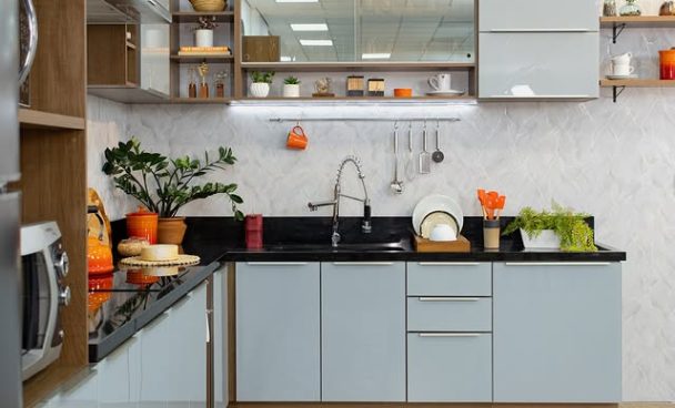 Cozinha com armários cinza-claro e detalhes em madeira, bancada preta, pia de aço inox, prateleiras abertas com utensílios e plantas, decorada com diversos objetos laranja.