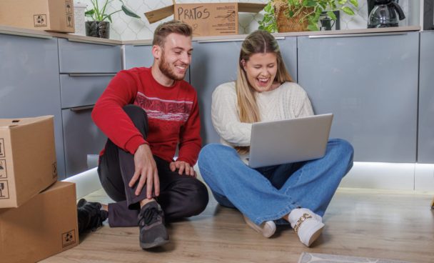 Casal sentado no chão de uma cozinha enquanto olham oportunidades de móveis na black friday no notebook. A cozinha conta com armários Madesa cinza e caixas de mudança