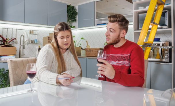 um homem e uma mulher loiros e de pele branca estão sentados em uma bancada no centro de uma cozinha gourmet. Ao fundo está uma cozinha Madesa e caixas de mudança, assim como eletrodomésticos dispostos sobre os balcões. A mulher veste um cardigã branco e o homem um vermelho, e ambos estão bebendo vinho em taças de vidro enquanto fazem anotações sobre a bancada da ilha.
