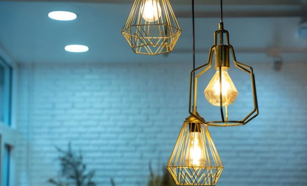 Três luminárias pendentes de metal dourado com lâmpadas expostas, iluminando um ambiente moderno com paredes de tijolos brancos. A luz suave cria uma atmosfera acolhedora e contemporânea.