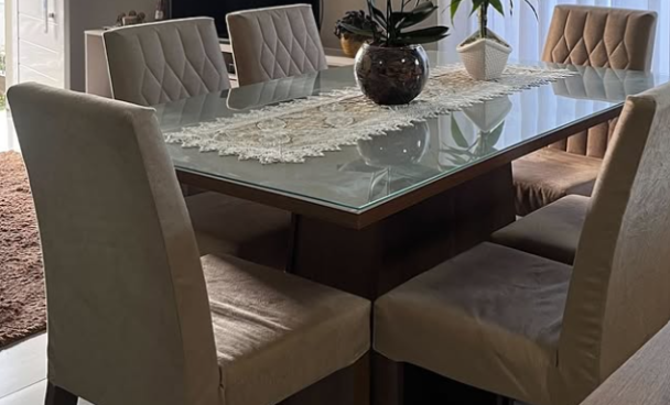 Mesa de jantar de vidro com seis cadeiras estofadas em tecido bege, decorada com um vaso de flores na sala de jantar, ao lado de cortinas brancas.