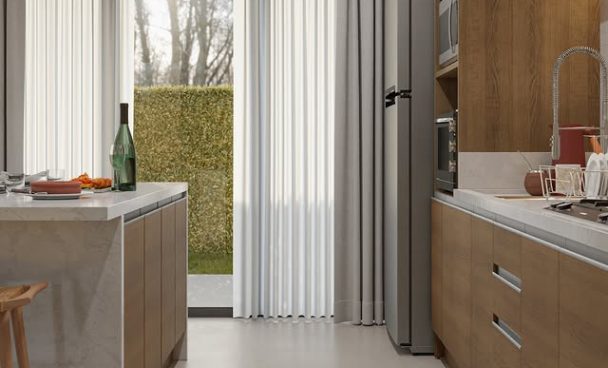 Cozinha com armários de madeira clara e branco, bancada de mármore, geladeira de inox, micro-ondas embutido, pia com torneira e cortinas brancas que cobrem uma grande janela. Há uma porta de vidro que leva ao jardim com árvores e arbustos ao fundo.