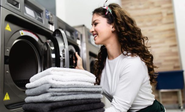 Mulher sorridente colocando toalhas em uma máquina de lavar, com pilha de toalhas ao lado.