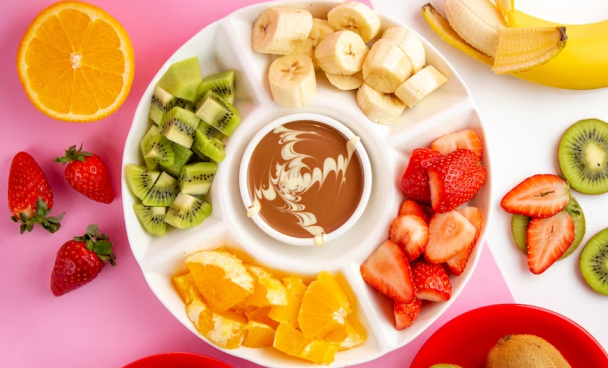 A imagem ilustra um conjunto de frutas para fondue com chocolate ao centro. Elas estão em uma bandeja branca e o fundo da foto é rosa.