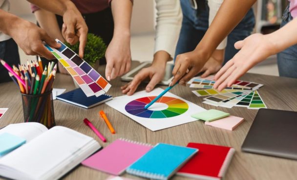 as mãos de diversas pessoas estão sobre uma mesa com cartilhas de cores e um círculo cromático impresso. Alguns deles seguram algumas cartelas de cores na mão enquanto outros seguram canetas e lápis, apontando para o círculo cromático na hora de fazer combinações.