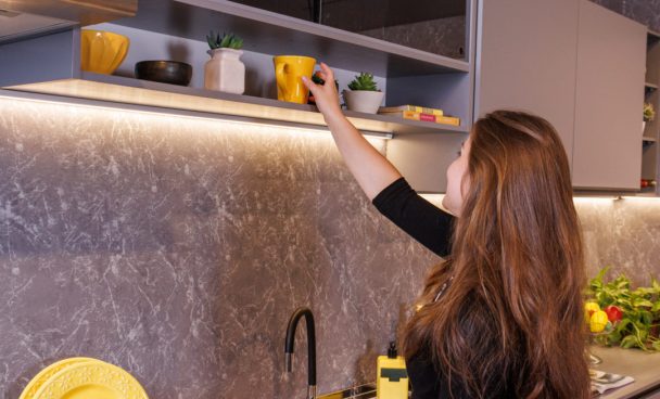Mulher jovem, esticando o braço para guardar louças num nicho de armário aéreo da cozinha Nice Madesa.