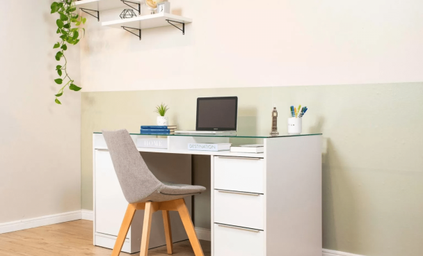 uma sala clara com uma parede pintada em divisões horizontais entre branco e verde claro está em foco. Ela conta com uma escrivaninha branca com gavetas e um notebook sobre seu tampo de vidro. Uma cadeira com pés de madeira está em frente à escrivaninha e, na parede, duas prateleiras estão segurando vasos e objetos decorativos.