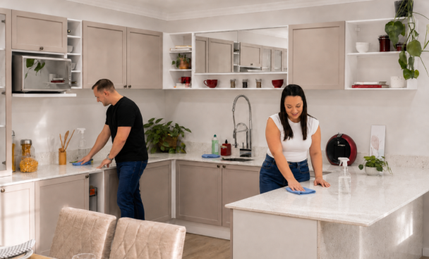 Casal na faxina pesada da cozinha em L bege, limpando a bancada antes de receber visita