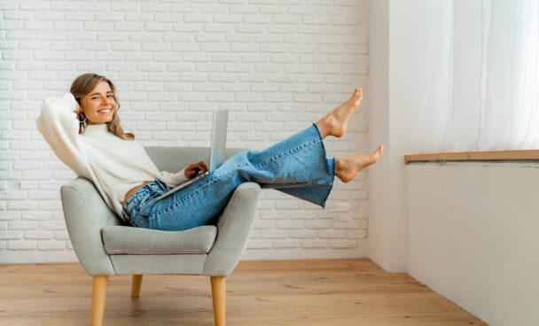Smiling businesswoman sitting on sofa at home working on laptop computer.   Wearing cozy sweater.