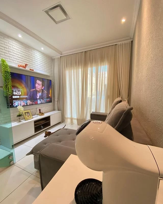 A imagem ilustra uma sala de estar com uma tv instalada em cima de um hack branco, representando um presente para casa nova.