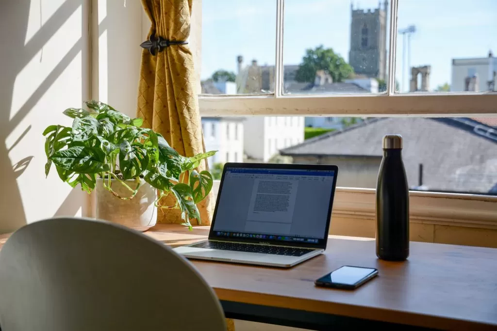 Home office no quarto montado em frente a uma janela, com notebook, celular, garrafa d'água e decorado com uma planta.