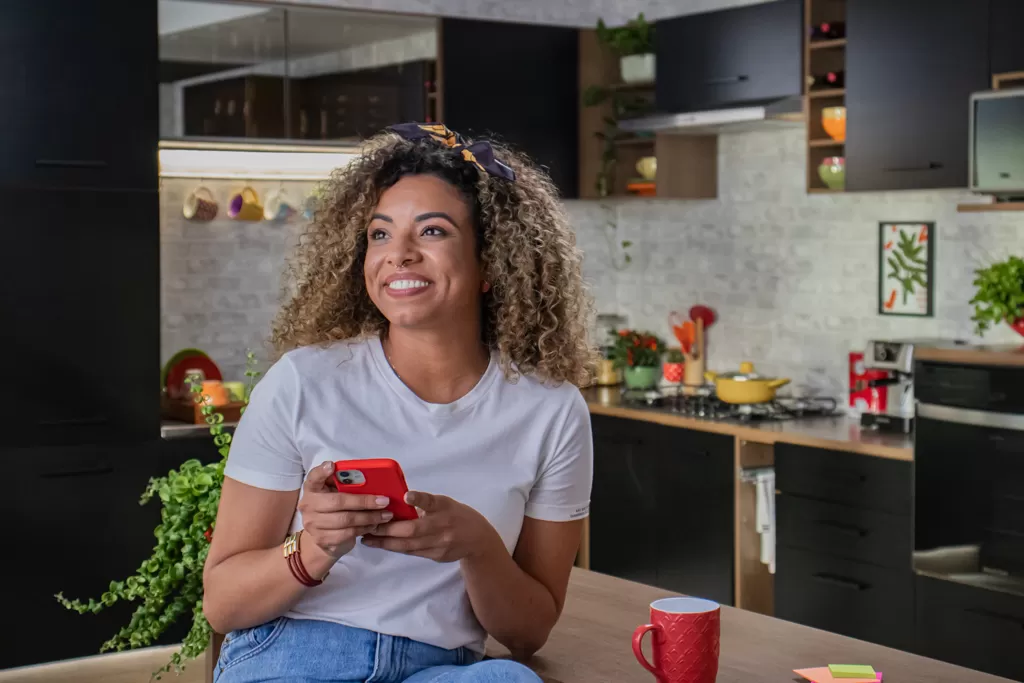 Mulher negra, sorrindo, sentada numa bancada enquanto segura um smartphone.