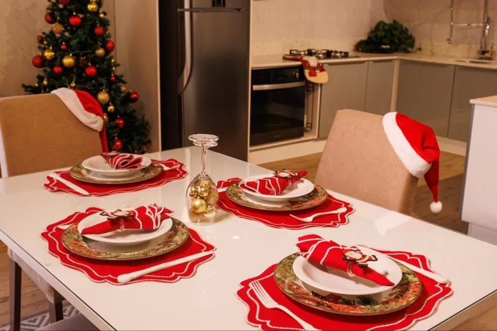 Mesa posta de Natal Madesa, decorada com sousplat vermelho e decorações douradas. Ao fundo, uma cozinha Madesa decorada e uma árvore de Natal.