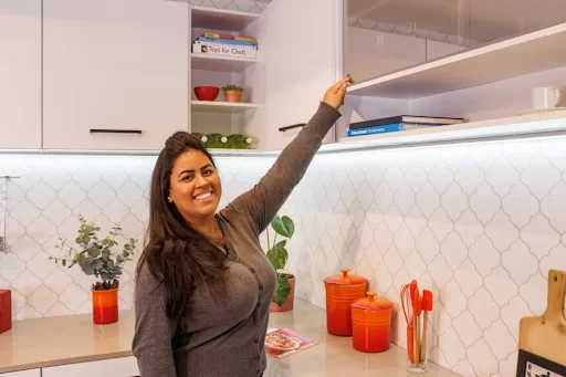 Mulher sorrindo, enquanto estica o braço para alcançar o armário aéreo na cozinha.