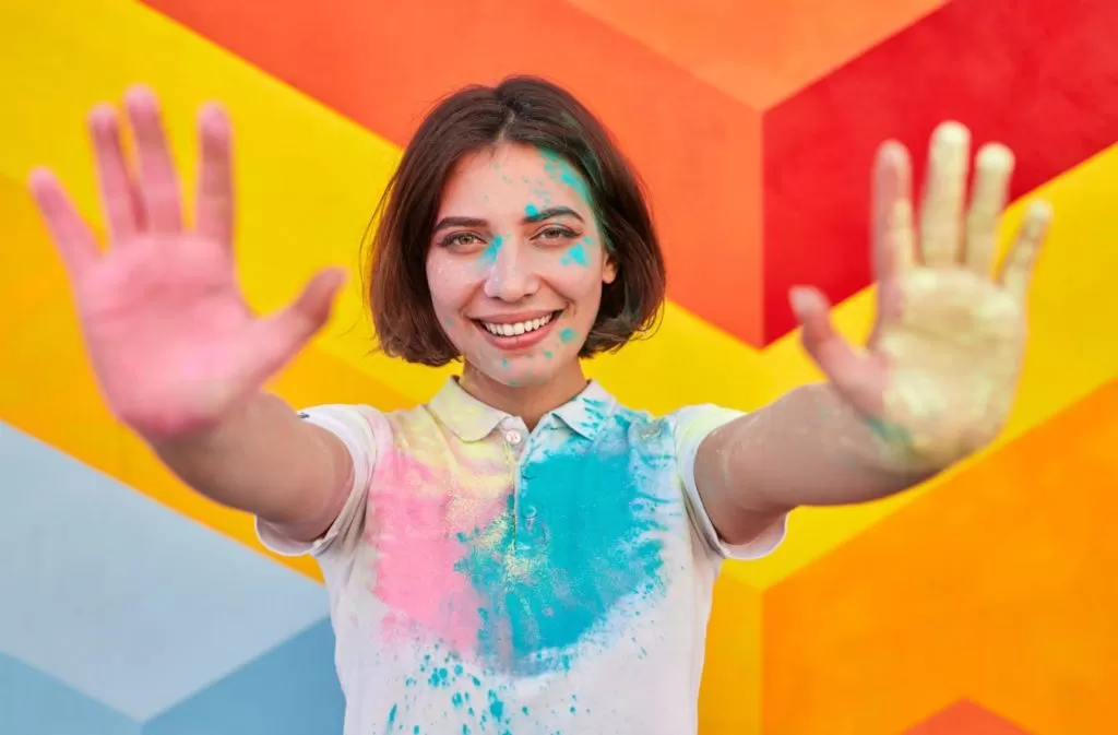 A imagem ilustra uma jovem de cabelo curto chanel em uma parede colorida e com as mãos pintadas de tinta simbolizando a pintura geométrica.