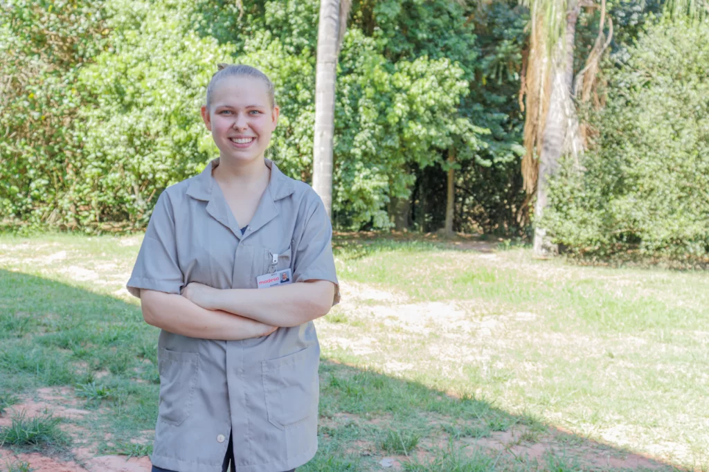 A imagem ilustra uma colaboradora madesa em um cenário ao ar livre. Ela é loira, branca, e veste um uniforme cinza, representando as mulheres no mercado de trabalho.