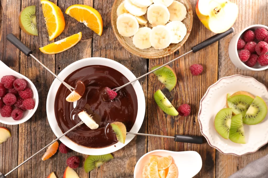 A imagem ilustra um conjunto de frutas para fondue colocadas numa mesa ao redor do rechaud.