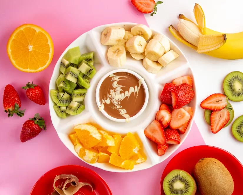 A imagem ilustra um conjunto de frutas para fondue com chocolate ao centro. Elas estão em uma bandeja branca e o fundo da foto é rosa.