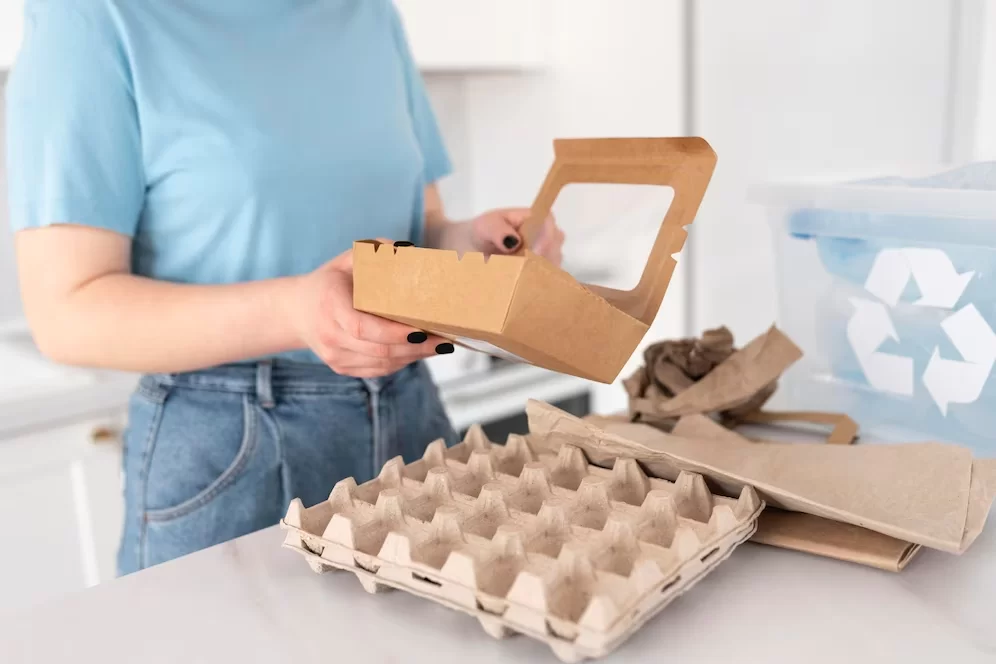 A imagem ilustra ideias de reciclagem com uma mulher segurando embalagens de papelão. Ela usa uma blusa azul e calça jeans.