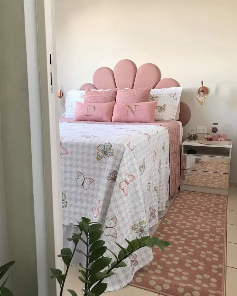 A imagem ilustra um quarto com uma cama de casal com colcha florida em branco e almofadas e cabeceira rosa claro. O quarto possui mesa de cabeceira e detalhes decorativos em cores que combinam com rosa.