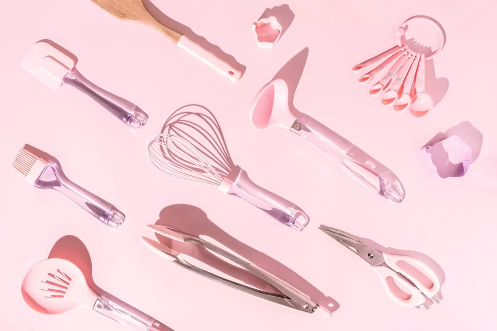 A imagem ilustra utensílios de cozinha como pegador, batedor de ovos e descascador na cor rosa. 