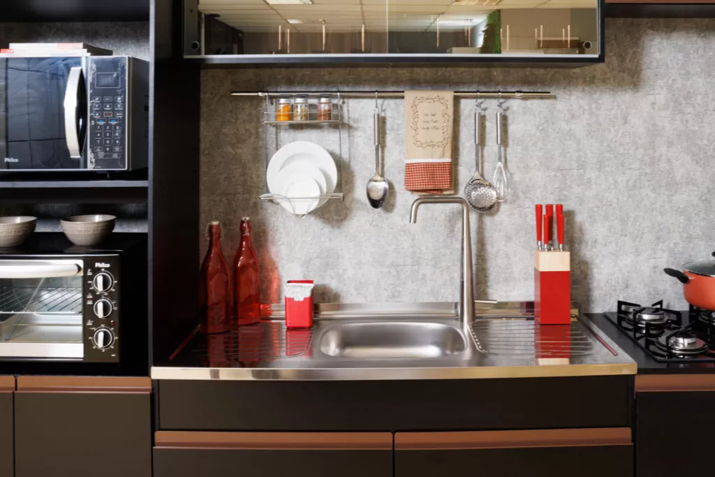 Cozinha americana compacta com pia em aço inox e torneira de aço. Em cima há garrafas de vidro vermelhas com utensílios também vermelhos. Em cima um escorredor de louça suspenso com pratos brancos. Ao lado um micro-ondas e um forno elétrico.