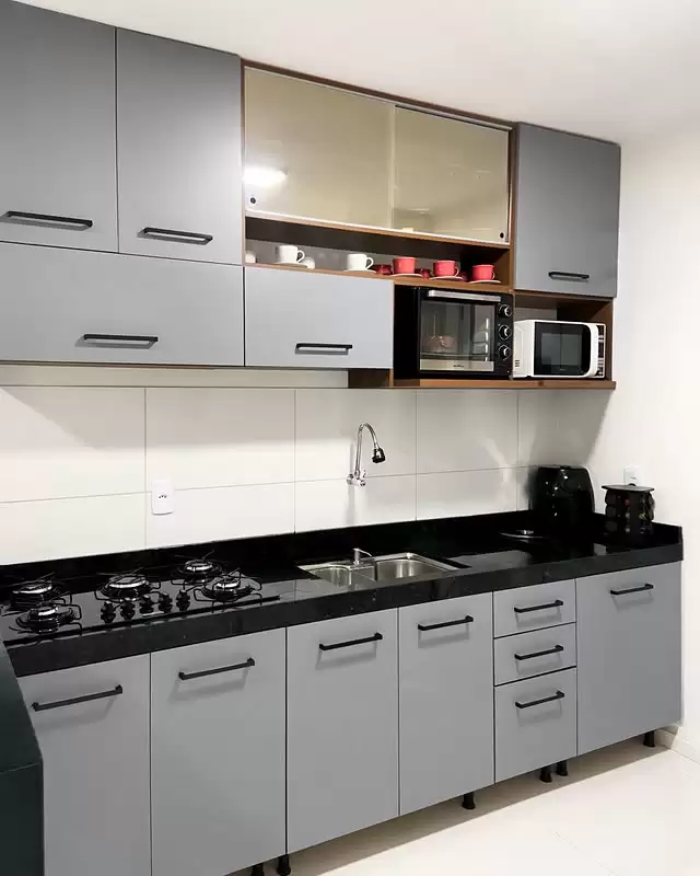 Cozinha com armários superiores e inferiores em tom cinza, um armário com porta de correr, bancada preta, pia de aço inox, micro-ondas e forno elétrico embutidos na parte superior, e utensílios de cozinha na bancada, com parede clara ao fundo.