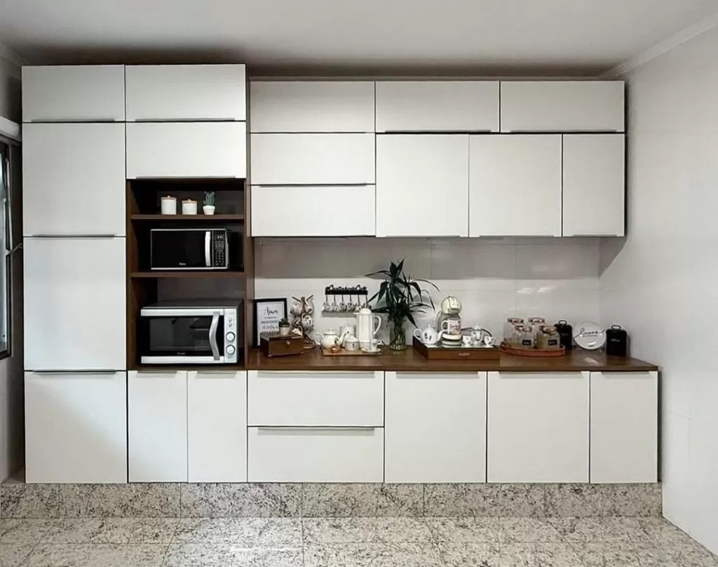 Cozinha com armários brancos, incluindo uma área central com nicho de madeira que contém micro-ondas e outros eletrodomésticos, e uma bancada de madeira com utensílios, plantas e itens decorativos.