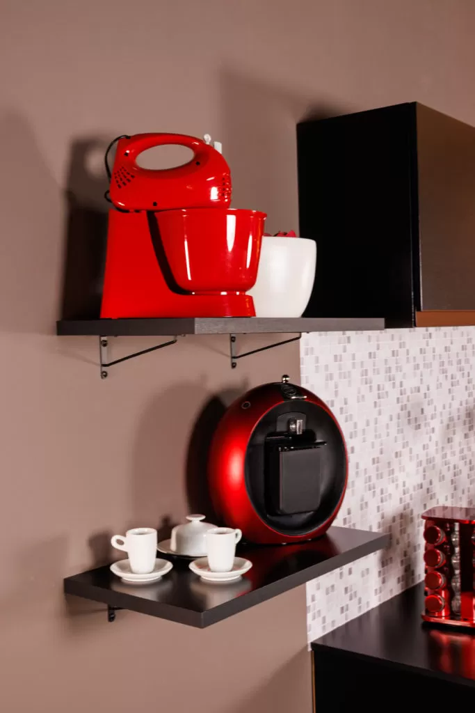 Duas prateleiras suspensas instaladas na parede de uma cozinha, uma com uma máquina de café expresso vermelha e xícaras com pires brancos ao lado, e outra com uma batedeira vermelha e uma tigela branca, com parede de azulejos pequenos ao fundo.
