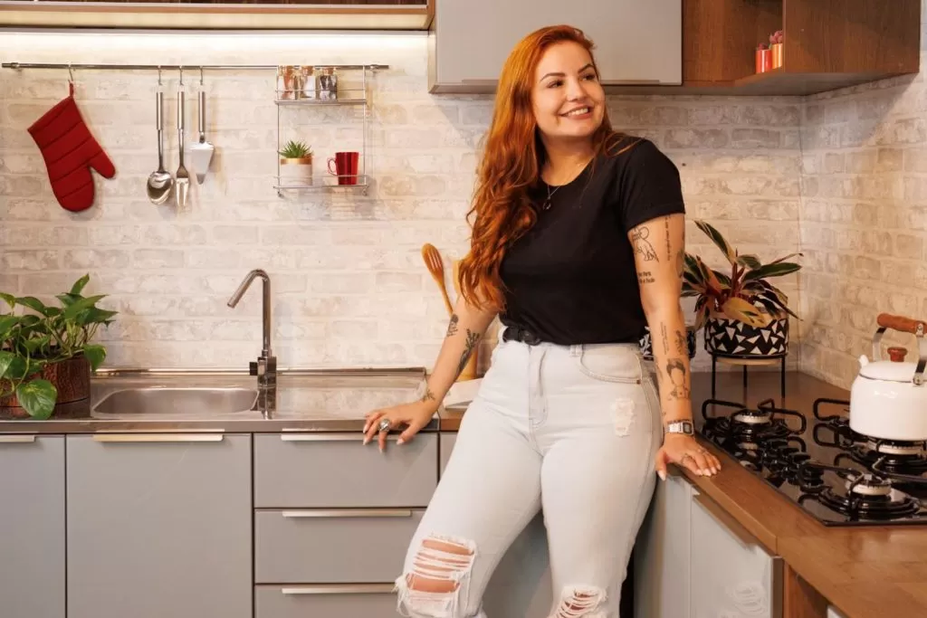 Mulher ruiva, sorrindo, posando em frente à cozinha Lux, na cor cinza e amadeirado escuro.