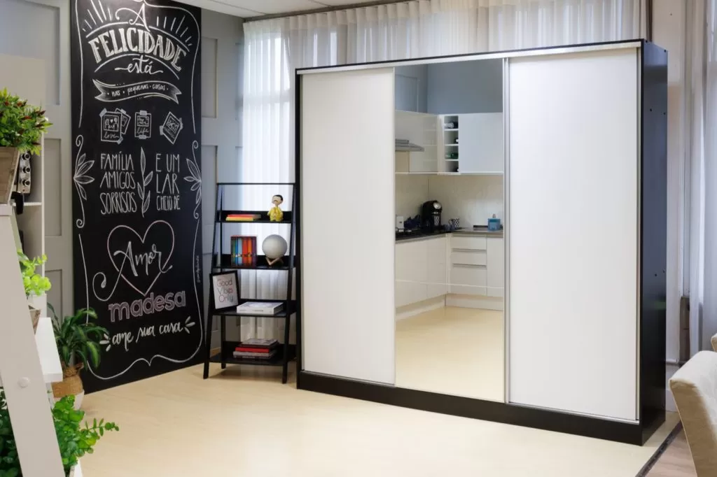Guarda-roupa Royale, na cor branca, ao lado de uma estante escada decorada e um quadro negro com desenhos sobre família, felicidade e os escritos “Madesa, ame sua casa”.